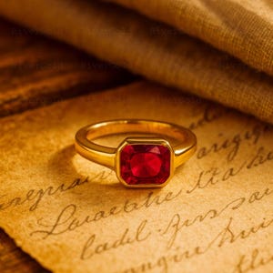 Może przedstawiać: Złoty pierścionek z kwadratowym, żywym, czerwonym kamieniem szlachetnym. Pierścionek leży na postarzanym papierze z odręcznym pismem. Tło obejmuje drapowaną, teksturowaną tkaninę.