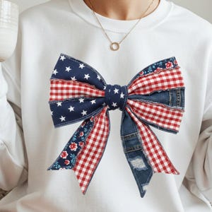 Pode incluir: Camisola branca com um grande design de laço. O laço incorpora ganga, xadrez vermelho, floral e tecido azul marinho com estrelas brancas. O design tem um tema patriótico, com o texto "PNG-300 DPI" no canto superior direito.