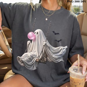 May include: A charcoal gray t-shirt featuring a graphic of a white ghost sitting on a wooden swing, blowing a pink bubble gum bubble. Two small black bats are flying to the right of the ghost. The shirt has a crew neckline and short sleeves.