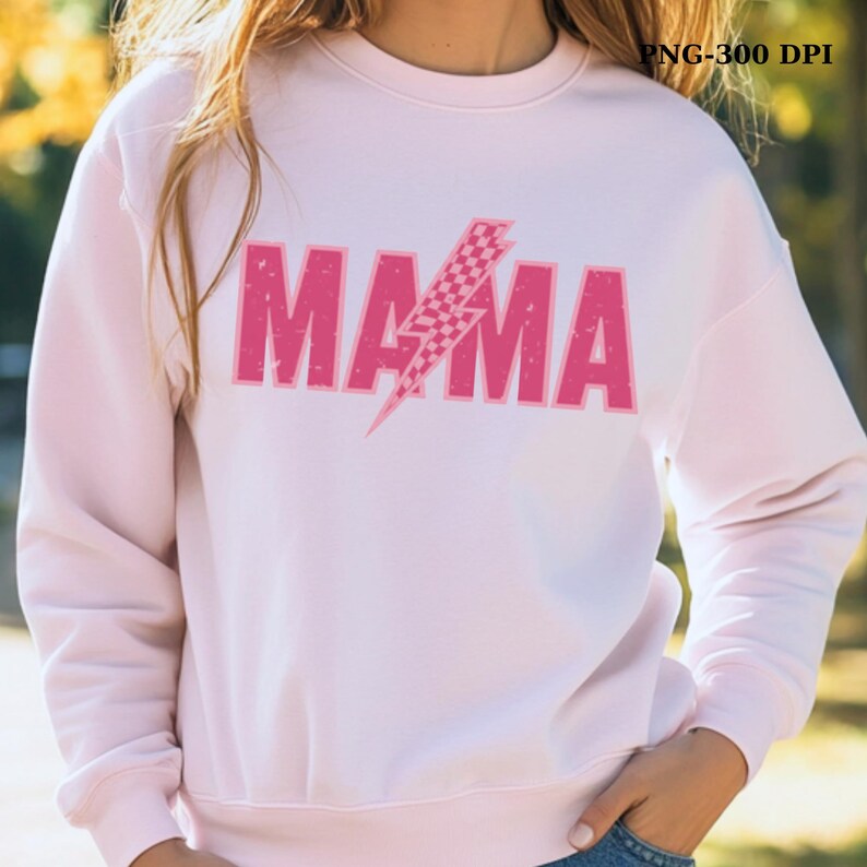 May include: A light pink sweatshirt with the word "MAMA" in distressed pink block letters. A pink and white checkered lightning bolt graphic is centered over the word. The sweatshirt has a crew neck and long sleeves.