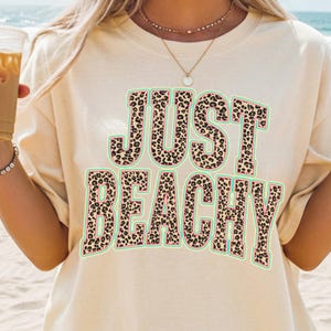 以下が含まれることがあります： 「JUST BEACHY」の文字がヒョウ柄でデザインされた、クリーム色のTシャツ。 ターコイズの縁取りとピンクのボーダーが特徴です。カジュアルなビーチをテーマにしたアパレルアイテムです。