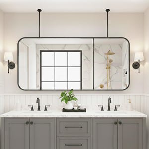 Op de afbeelding: Een moderne badkamer met een grote, rechthoekige spiegel met zwart frame die aan het plafond hangt. De wastafel heeft een grijze kast met twee wastafels, zwarte kranen en wandlampen. Een plant en decoratieve items staan op het aanrecht.