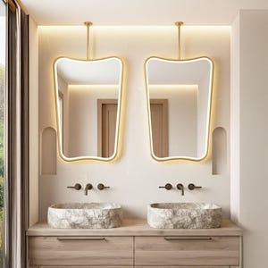 Può includere: Un moderno mobile da bagno con due lavabi in pietra e specchi con cornice dorata abbinati. Gli specchi hanno una forma unica e sono sospesi. Il mobile ha armadi in legno chiaro e rubinetti neri.