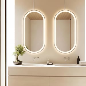 Può includere: Due specchi ovali illuminati sono appesi sopra un moderno mobile da bagno. Gli specchi hanno un contorno luminoso e morbido. Un piccolo bonsai è appoggiato sul piano di lavoro. Il mobile è di un colore neutro chiaro, con un design minimalista.