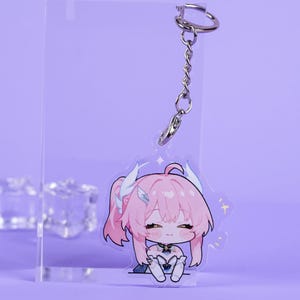 May include: A clear acrylic keychain featuring a cartoon character with pink hair and a silver keychain attachment. The character is sitting with a relaxed expression. The background is a solid lavender color.