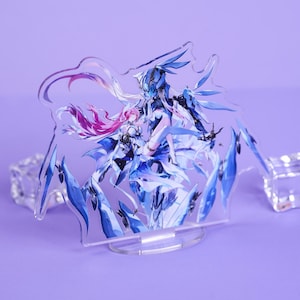 May include: A clear acrylic stand featuring a detailed illustration of two anime-style characters in shades of blue, white, and pink. The artwork is mounted on a clear base, with two clear cube-shaped accents on either side. The background is a solid lavender color.