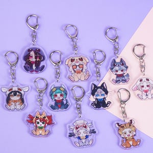 K&ouml;nnte beinhalten: Eine Sammlung von Acryl-Schl&uuml;sselanh&auml;ngern mit Anime-Charakteren in verschiedenen Outfits. Jeder Schl&uuml;sselanh&auml;nger hat einen silberfarbenen Metallring und -verschluss. Die Charaktere sind in leuchtenden Farben vor einem lila und rosa Hintergrund dargestellt.