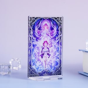 May include: A clear acrylic stand featuring a vibrant illustration of two anime-style figures in shades of purple, blue, and white. The artwork is framed by a decorative border. The stand is on a light blue surface.