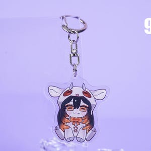 K&ouml;nnte beinhalten: Ein klarer Acryl-Schl&uuml;sselanh&auml;nger mit einer Cartoonfigur in einem Kuhkost&uuml;m. Die Figur hat schwarzes Haar, eine Brille und eine orangefarbene Schleife. Der Schl&uuml;sselanh&auml;nger ist an einem silbernen Metallverschluss befestigt. Die Zahl 9 befindet sich in der oberen rechten Ecke.