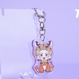 K&ouml;nnte beinhalten: Ein klarer Acryl-Schl&uuml;sselanh&auml;nger mit einer Cartoonfigur in einem Tigerkost&uuml;m. Die Figur hat orange-braunes Fell, eine Fliege und sitzt. Der Schl&uuml;sselanh&auml;nger hat einen silbernen Verschluss und einen silbernen Ring. Der Hintergrund ist helllila.