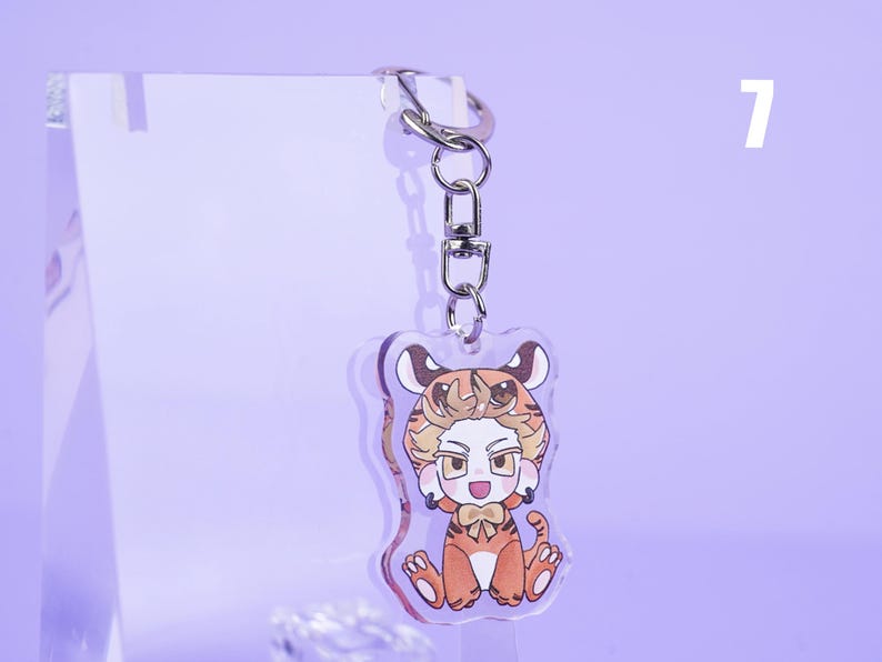 K&ouml;nnte beinhalten: Ein klarer Acryl-Schl&uuml;sselanh&auml;nger mit einer Cartoonfigur in einem Tigerkost&uuml;m. Die Figur hat orange-braunes Fell, eine Fliege und sitzt. Der Schl&uuml;sselanh&auml;nger hat einen silbernen Verschluss und einen silbernen Ring. Der Hintergrund ist helllila.