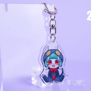 K&ouml;nnte beinhalten: Ein klarer Acryl-Schl&uuml;sselanh&auml;nger mit einer Cartoonfigur in einem dunkelblauen Kapuzen-Outfit mit wei&szlig;en H&ouml;rnern und einer roten Fliege. Die Figur hat t&uuml;rkisfarbenes Haar und rote Augen. Der Schl&uuml;sselanh&auml;nger ist an einem silbernen Metallring befestigt.