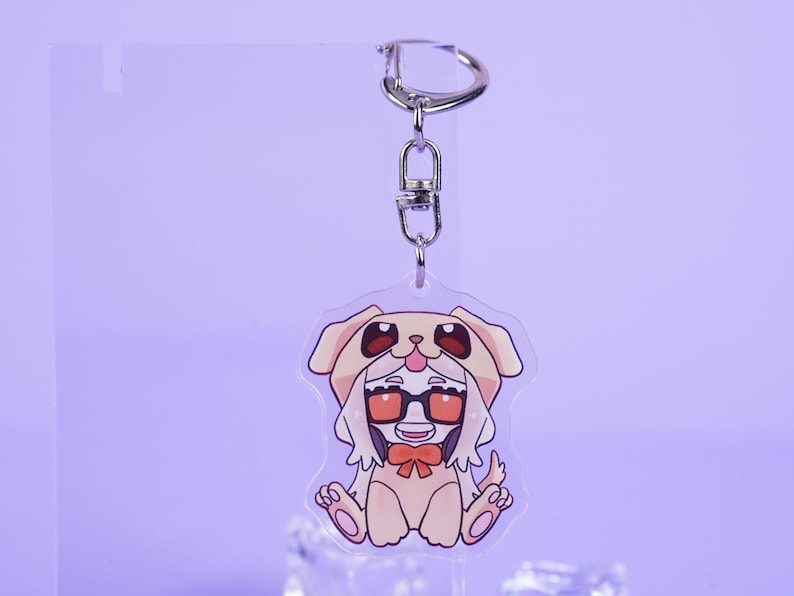 K&ouml;nnte beinhalten: Ein klarer Acryl-Schl&uuml;sselanh&auml;nger mit einer Zeichentrickfigur in einem Hundekost&uuml;m mit Brille und Fliege. Der Schl&uuml;sselanh&auml;nger hat einen silbernen Metallverschluss und -ring. Die Figur ist in den Farbt&ouml;nen Beige, Rot und Schwarz gehalten.