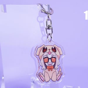 K&ouml;nnte beinhalten: Ein klarer Acryl-Schl&uuml;sselanh&auml;nger mit einer Cartoon-Figur in einem Hundekost&uuml;m. Die Figur tr&auml;gt eine Brille und eine Fliege. Der Schl&uuml;sselanh&auml;nger ist an einem silbernen Metallverschluss und -ring befestigt. Die Zahl 10 befindet sich im Hintergrund.