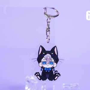 K&ouml;nnte beinhalten: Ein klarer Acryl-Schl&uuml;sselanh&auml;nger mit einer Cartoonfigur in einem schwarzen Katzenkost&uuml;m mit einer blauen Schleife. Der Schl&uuml;sselanh&auml;nger hat einen silbernen Metallverschluss und -ring. Die Nummer 6 ist im Hintergrund sichtbar.