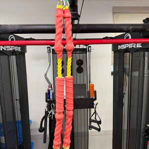 May include: Red and yellow exercise resistance bands hanging from a black and red Inspire fitness machine. The bands have a twisted design and are attached with metal carabiner clips. The background includes other gym equipment.