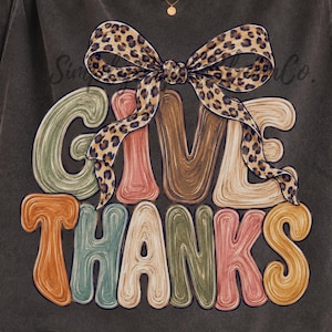 May include: A charcoal gray short-sleeve t-shirt featuring the text GIVE THANKS in a multi-colored, wavy, retro-style font. A large leopard print bow is positioned above the text. The shirt has a faded, vintage-inspired wash and crew neckline.
