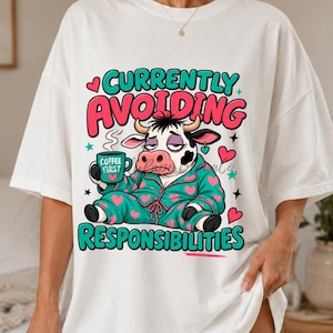 May include: A white t-shirt featuring a graphic of a cow wearing teal pajamas with pink hearts, holding a teal mug labeled COFFEE FIRST. The text reads CURRENTLY AVOIDING RESPONSIBILITIES in teal and pink, surrounded by small pink hearts and stars.