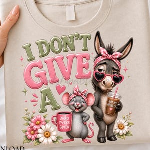 May include: A beige crewneck sweatshirt featuring a graphic of a donkey and a mouse wearing pink headbands. The donkey wears heart-shaped sunglasses and holds a coffee cup labeled NOT MY PROBLEM. The mouse holds a mug labeled ZERO F*CKS GIVEN.