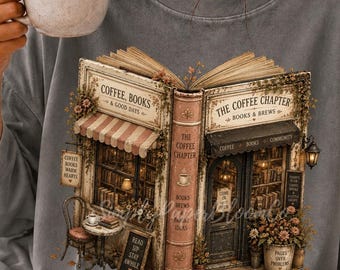 May include: A charcoal gray t-shirt featuring a graphic print of an open book that transforms into a cozy coffee shop. The illustration includes text reading Coffee, Books & Good Days and The Coffee Chapter, with detailed storefront elements.