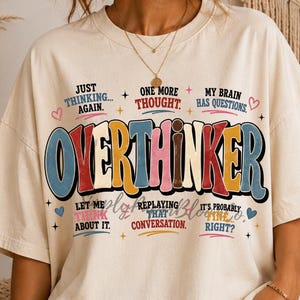 May include: A cream-colored short-sleeve t-shirt featuring the word OVERTHINKER in large, multi-colored retro-style lettering. The design includes smaller text phrases like JUST THINKING AGAIN, ONE MORE THOUGHT, and MY BRAIN HAS QUESTIONS.