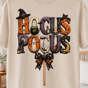 May include: A cream-colored t-shirt featuring a graphic print of the text HOCUS POCUS in large, stylized letters. Each letter contains Halloween-themed illustrations including a witch hat, ghost, black cat, broom, and lantern, finished with a bow.