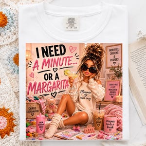 May include: A white t-shirt featuring a graphic illustration of a person holding a margarita with the text I NEED A MINUTE... OR A MARGARITA. The design includes various items like a coffee cup, a pink mug, a teddy bear, and small toys.