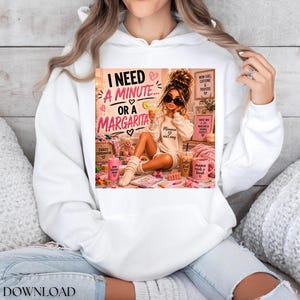 May include: A white hooded sweatshirt featuring a graphic print of a person with sunglasses holding a drink, surrounded by text that reads I NEED A MINUTE OR A MARGARITA, along with various pink and brown accessories and small decorative items.