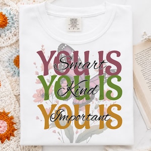 May include: White t-shirt with the words "You are Smart, Kind, Important" in burgundy, green, and gold. A butterfly graphic is centered on the shirt, with floral accents. The shirt is folded on a white surface.