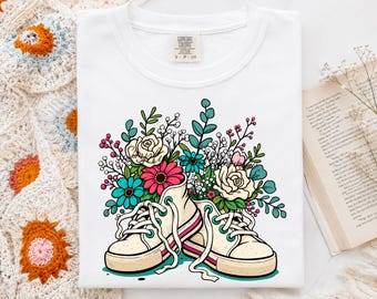 May include: A white t-shirt featuring a graphic illustration of two cream-colored canvas sneakers overflowing with a vibrant bouquet of pink, teal, and white flowers, green leaves, and small berries, with a Comfort Colors brand tag at the neckline.