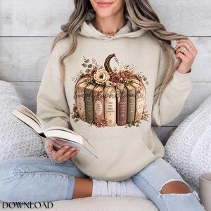 May include: A beige hooded sweatshirt featuring a graphic print of a pumpkin constructed from stacked books. The pumpkin is adorned with floral accents and the text SimplyPaperBloomCo. The books include phrases like Just One More Chapter.