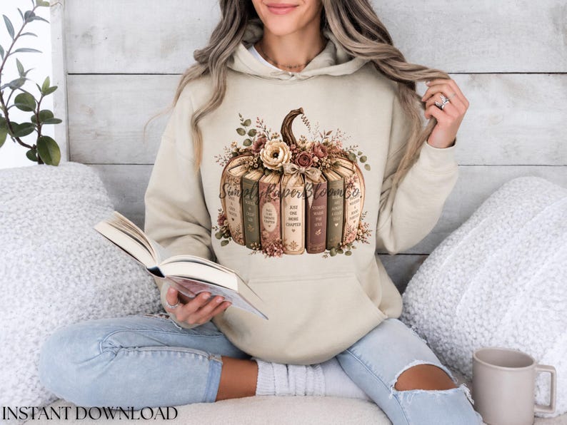May include: A beige hooded sweatshirt featuring a graphic print of a pumpkin constructed from stacked books. The pumpkin is adorned with floral accents and the text SimplyPaperBloomCo. The books include phrases like Just One More Chapter.