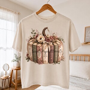 May include: A cream-colored short-sleeve t-shirt featuring a graphic of a pumpkin constructed from stacked books. The books are decorated with floral accents and include text on their spines. The design is centered on the front of the shirt.