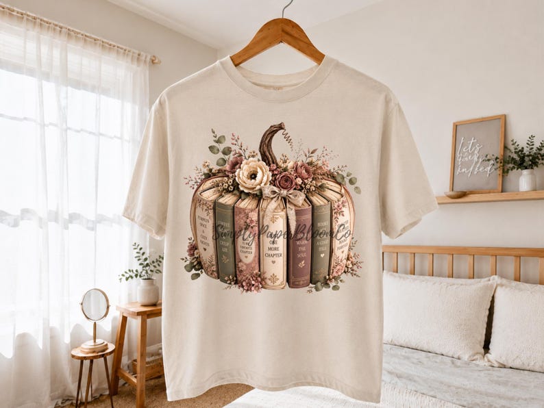 May include: A cream-colored short-sleeve t-shirt featuring a graphic of a pumpkin constructed from stacked books. The books are decorated with floral accents and include text on their spines. The design is centered on the front of the shirt.