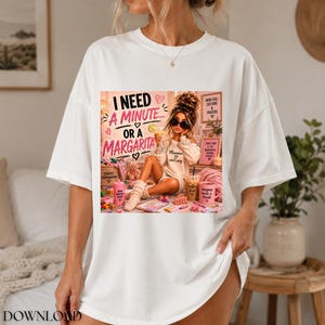 May include: A white short-sleeved t-shirt featuring a graphic print of a person with sunglasses sitting amidst various items, including a drink and a pink mug. The text reads: I NEED A MINUTE... OR A MARGARITA, MOM FUEL, CAFFEINE & PRAYERS, and more.