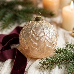 Peut inclure: Une boule de Noël sphérique en verre couleur champagne avec un motif en chevrons texturé en relief. La partie supérieure est ornée de paillettes dorées, de petites perles blanches et de sequins, avec une attache dorée.