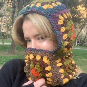 May include: A colorful crochet balaclava in a granny square pattern. The balaclava features a mix of colors including yellow, brown, green, orange, purple, and blue. The balaclava covers the head and neck, with an opening for the face. The person is wearing a black sweater.