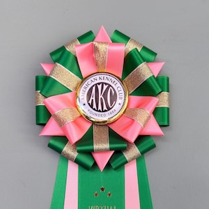 May include: A rosette ribbon with green, pink, and gold accents. The center features an "AKC" emblem. The ribbon reads "VIRTUAL SCENT WORK WINNER" in gold lettering. This award is for dog show competitions.