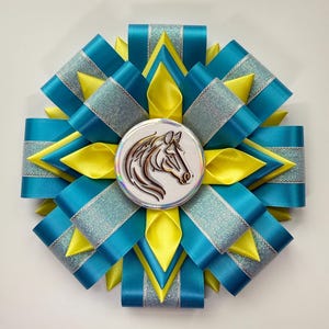 May include: A decorative rosette featuring a horse head emblem. The rosette is made of teal, yellow, and silver ribbons arranged in a starburst pattern. The center medallion has a detailed horse profile.
