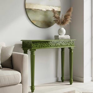 May include: A rectangular olive green console table with intricate carved lattice detailing along the front apron and four slender, turned legs. A white ceramic vase containing dried brown pampas grass sits on top of the table.