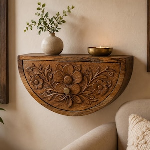 May include: A semi-circular wall-mounted wooden shelf featuring a carved floral pattern on the front drawer panel, a small round knob, and a flat top surface holding a light-coloured ceramic vase with greenery and a small metallic bowl with a candle.
