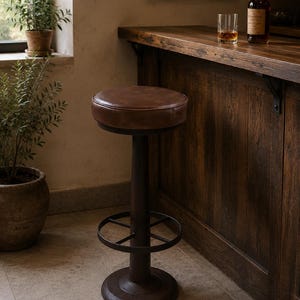 May include: A bar stool featuring a round, brown leather-upholstered seat atop a dark brown metal pedestal base. The base includes a circular footrest ring and a wide, flat circular floor plate, providing a stable foundation for the seat.