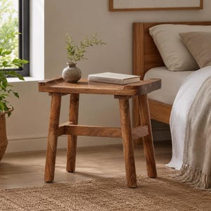 May include: A small, rectangular wooden side table with four sturdy, slightly angled legs and a thick top surface. A small, round, textured grey ceramic vase containing green sprigs and a white book are placed on the table's surface.