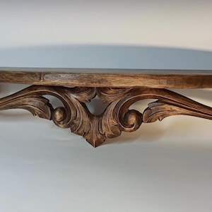Peut inclure: Étagère en bois brun foncé ornée d'un motif sculpté et enroulé. L'étagère est rectangulaire avec un bord avant incurvé et des supports décoratifs. L'étagère mesure environ 122 cm de long.