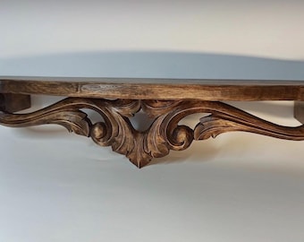 May include: A dark brown wooden wall shelf featuring a flat top surface and an ornate, carved support bracket underneath. The support displays intricate, symmetrical scrolling leaf patterns and curved decorative elements in a rich wood grain finish.