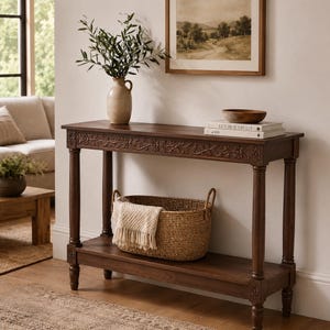 May include: A rectangular dark brown wooden console table featuring a carved floral pattern along the front apron and four turned legs. The table includes a lower shelf holding a woven wicker basket with a cream-coloured fringed textile inside.