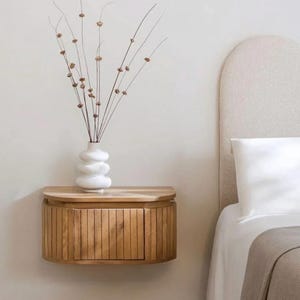 Könnte beinhalten: Ein hölzerner, an der Wand befestigter Nachttisch mit einer gebogenen Front und einer einzigen Tür. Eine weiße, skulpturale Vase hält getrocknete Stiele. Der Nachttisch ist aus hellem Holz gefertigt und an einer neutralfarbenen Wand befestigt.