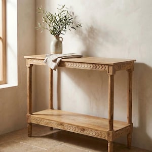 Peut inclure: Une console en bois brun clair avec une étagère inférieure et des détails décoratifs sculptés. La table a quatre pieds cylindriques et un plateau rectangulaire. Un vase avec des branches vertes et un tissu plié sont posés dessus.