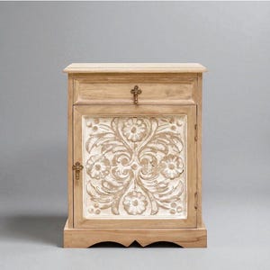 May include: A light brown wooden bedside table featuring a single top drawer with a small metal pull and a lower cabinet door. The door panel displays a carved floral pattern with white paint accents and a small metal latch handle on the side.