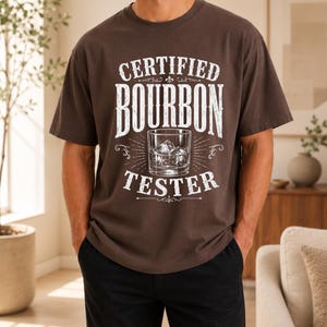 Peut inclure: Un t-shirt à manches courtes marron foncé avec une impression graphique blanche portant l'inscription CERTIFIED BOURBON TESTER. Le motif comprend une illustration centrale d'un verre rempli de liquide et de glaçons, entouré de lignes décoratives.